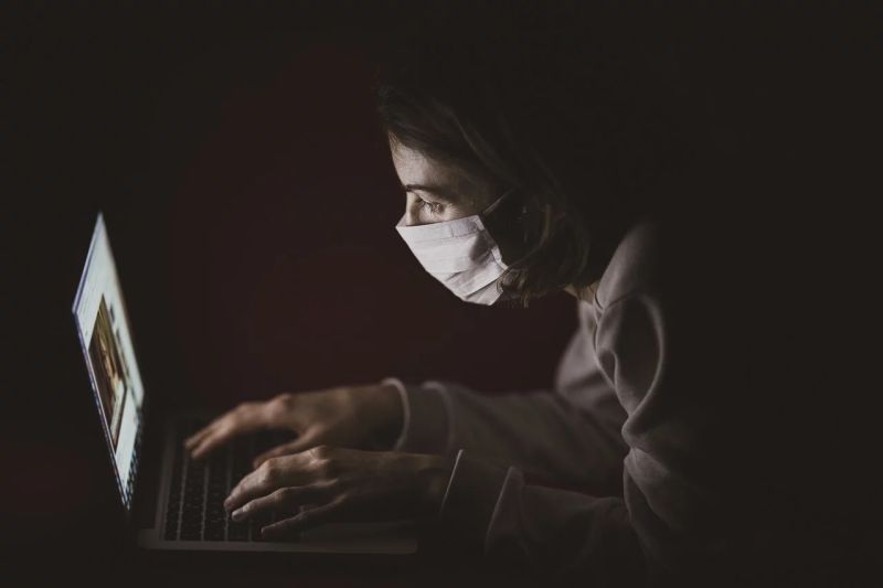 Memakai masker, seorang pria bekerja menggunakan laptopnya di sebuah kamar gelap. Foto Pixabay