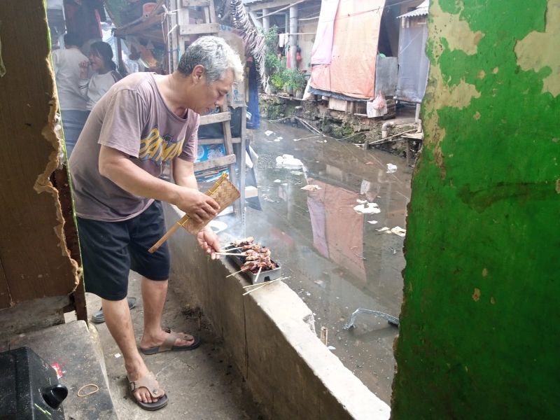 Seorang warga Jalan Jati Bunder, Tanah Abang, Jakarta Pusat, Ujang, tengah asyik membakar sate kambing di rumah kontrakannya yang berada di bantaran sungai penuh sampah, Minggu (10/7/2022). Alinea.id/Kudus Purnomo Wahidin.